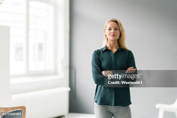 successful, blond businesswoman standing in bright room, with arms crossed - confidence stock-fotos und bilder