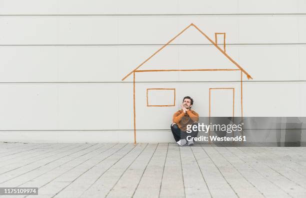 digital composite of young man sitting at a wall with a house - eigenheim deutschland stock-fotos und bilder