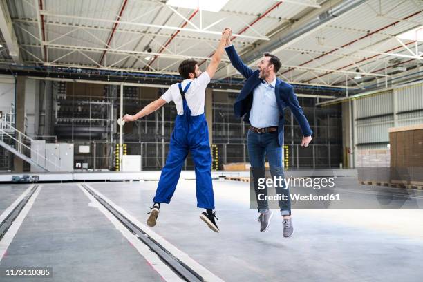 excited businessman and worker jumping and high fiving in a factory - workforce agility stock pictures, royalty-free photos & images