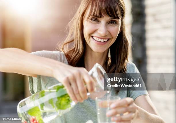 portrait of happy woman pouring infused water into glass - sunlight through drink glass foto e immagini stock