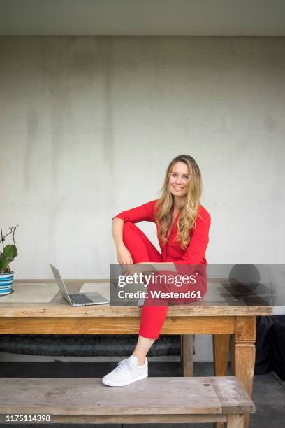 smiling woman with laptop sitting on wooden table at home - red jumpsuit stock pictures, royalty-free photos & images