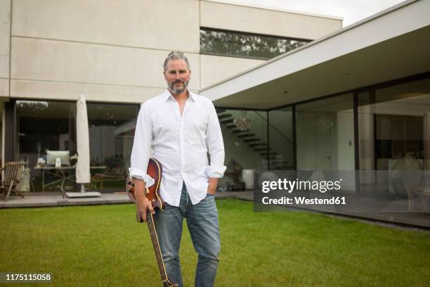 portrait of mature man standing on lawn in front of his house holding guitar - privilegien stil stock-fotos und bilder