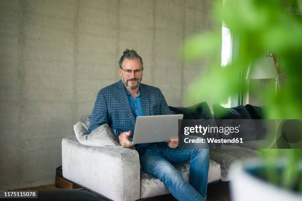 portrait of mature businessman sitting on couch at home using laptop - widerstand stock-fotos und bilder