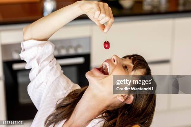 happy woman eating a cherry in kitchen at home - mouth open eating stock pictures, royalty-free photos & images