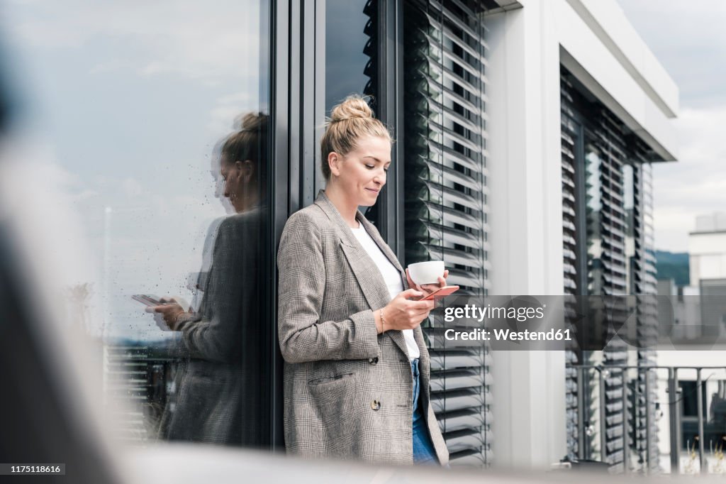 Businesswoman with cell phone and coffee cup standing on roof terrace