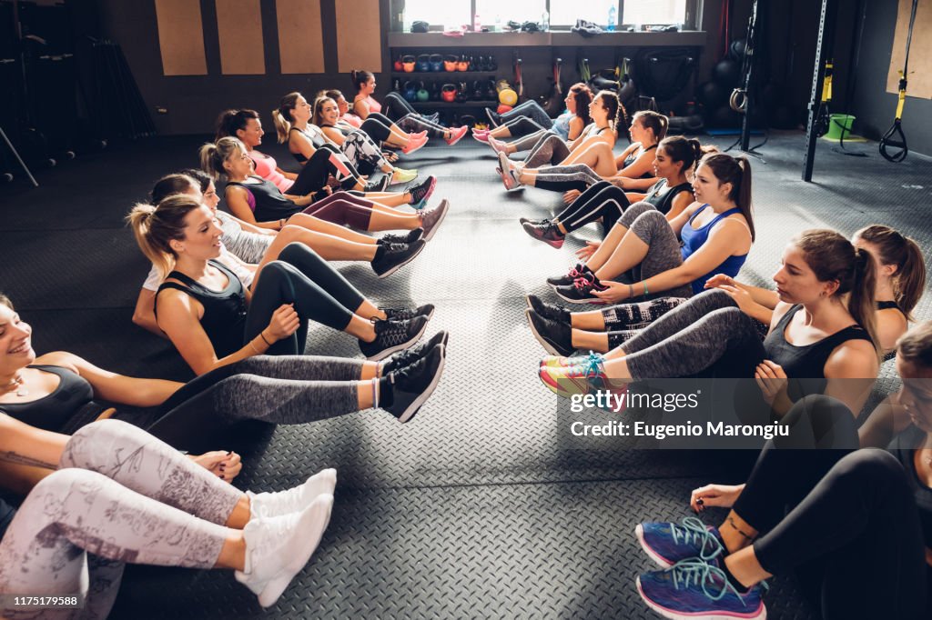 Large group of women training in gym, sitting in rows on floor with legs raised