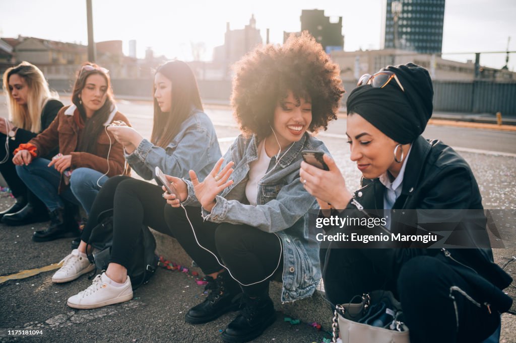 Friends sitting on kerb using smartphone, Milan, Italy