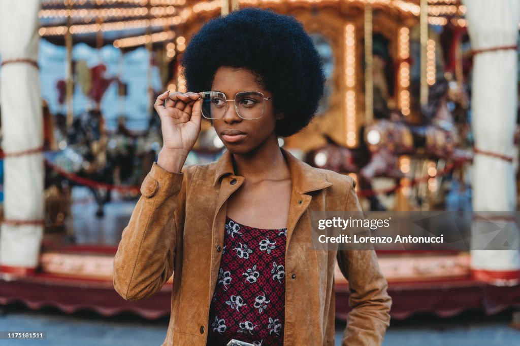 Young woman with afro hair putting on spectacles in front of carousel