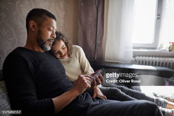 father and daughter sitting on couch at home using smartphone - father and daughter looking at smartphone together stock pictures, royalty-free photos & images