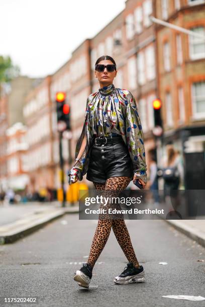 Giovanna Battaglia wears sunglasses, a shiny glitter lustrous turtleneck top with colored printed features, a belt, a black leather skirt, leopard...
