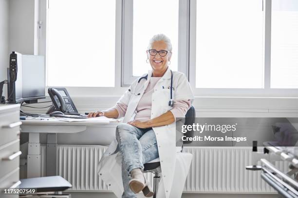 retrato de una doctora mayor sonriente en el escritorio - pies cruzados en los tobillos fotografías e imágenes de stock