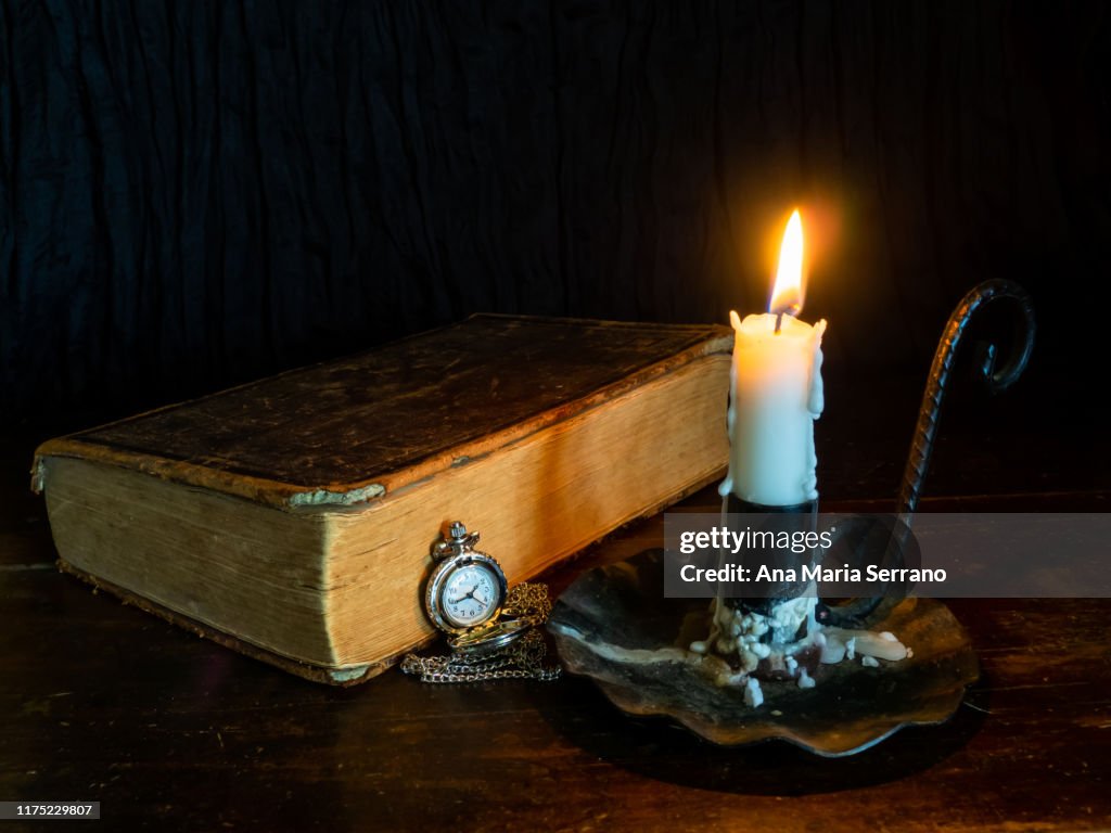 A still life with an ancient bible, a candlestick with a lit candle and a pocket chain watch that symbolize the passage of time