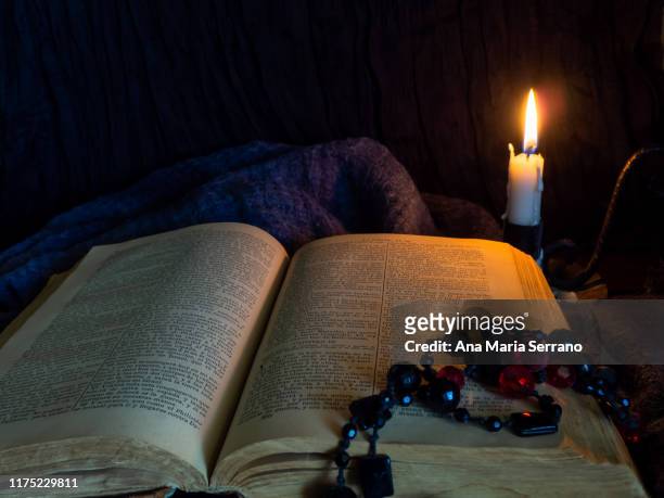 a still life with an open ancient bible, a candlestick with a lit candle and a rosary of black and red beads on a dark old cloth that symbolize the passage of time - sect stockfoto's en -beelden