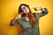 Young beautiful woman wearing green shirt and glasses over yelllow isolated background smiling cheerful showing and pointing with fingers teeth and mouth. Dental health concept.