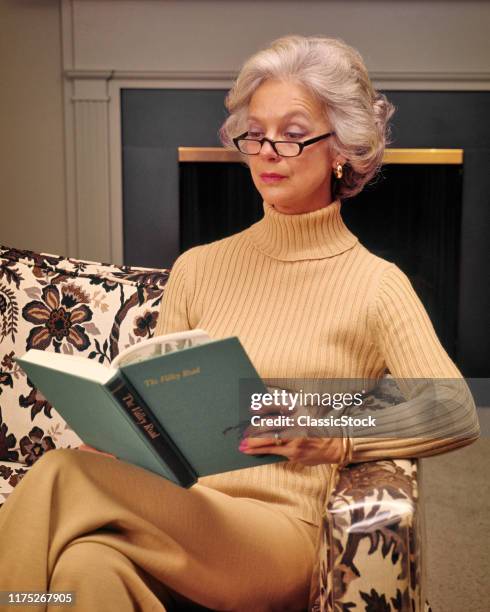 Serious Middle-Aged Gray Hair Woman Sitting On Living Room Sofa Wearing Turtleneck Reading Eyeglasses Enjoying A Book