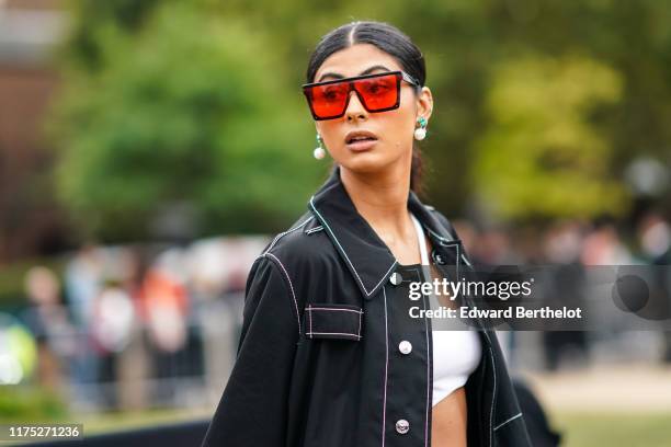 Guest wears red transparent sunglasses, a pearl earrings, a black open shirt with white stripes, white bras, during London Fashion Week September...