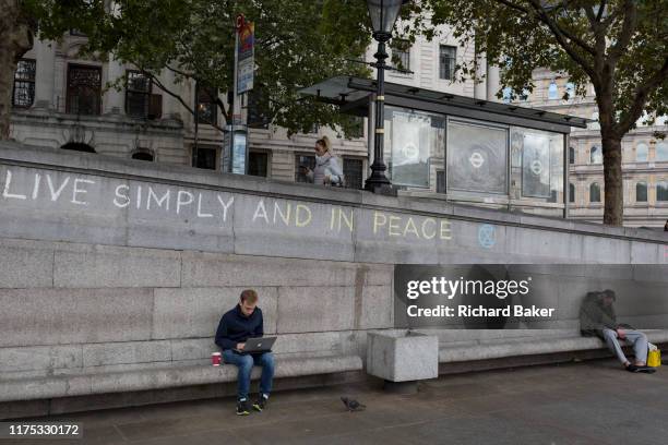 Man uses his laptop beneath the slogan 'Live Simply and Live in Peace' has been written on a wall by an environmental activist protesting about...