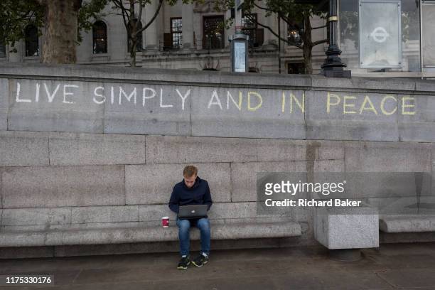 Man uses his laptop beneath the slogan 'Live Simply and Live in Peace' has been written on a wall by an environmental activist protesting about...