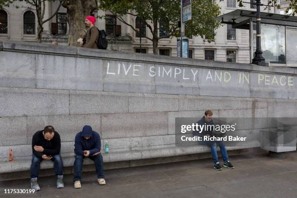 Three men use their phones and a laptop beneath the slogan 'Live Simply and Live in Peace' has been written on a wall by an environmental activist...