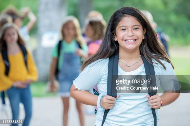 cute elementary age schoolgirl wearing backpack stands outside with group of friends - junior high student stock pictures, royalty-free photos & images