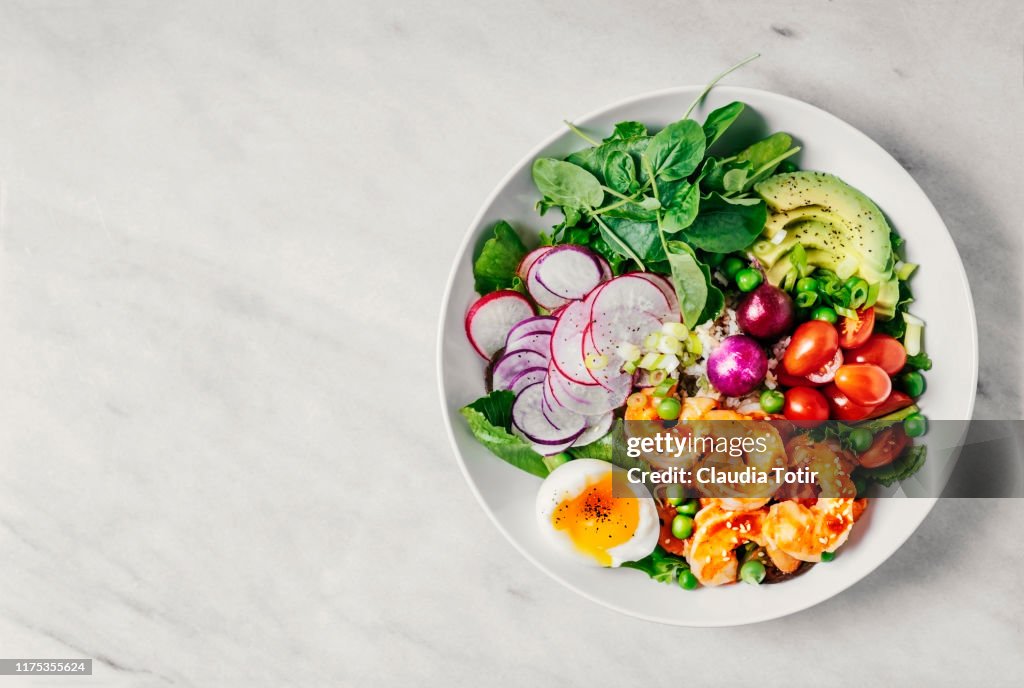 Healthy lunch bowl with greens, avocado, cherry tomatoes, radish, boiled egg, and shrimp on white background