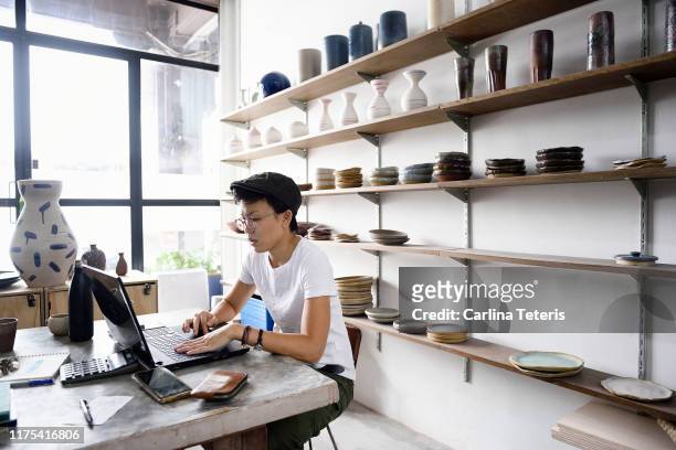 young asian entrepreneur working from her laptop in her store - small business stock pictures, royalty-free photos & images
