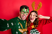 Photo of crazy couple making selfies sticking tongues winking eyes v-signing wear funky ugly ornaments jumpers isolated red color background