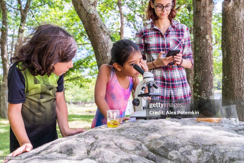 17-years-old teenager girl teaching her 9-years-old younger brother and 7-years-old sister how to working with a microscope and learning nature outdoors at the lakeshore in the sunny summer day.