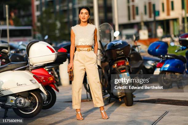 Camila Coelho wears a sequined shiny sleeveless cropped top, a belt, beige pants, a Vuitton clutch, silver shoes, outside the Alberta Ferretti show...