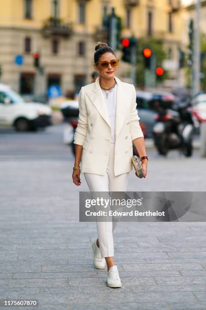 Olivia Palermo wears brown sunglasses, a white blazer jacket, a necklace, a white t-shirt, cropped pants, sneakers, a golden clutch, outside the...