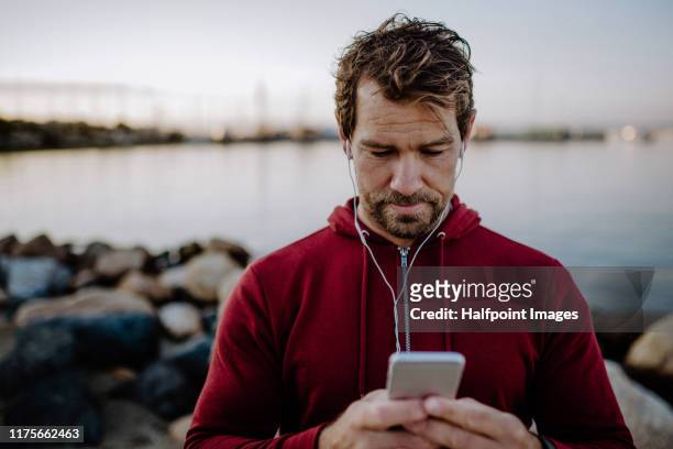 a fit mature sportsman runner with earphones standing outdoors on beach, using smartphone. - checking sports stock pictures, royalty-free photos & images