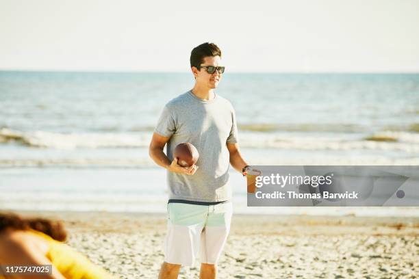 smiling man holding football and drink during party on beach - pantalón corto gris fotografías e imágenes de stock