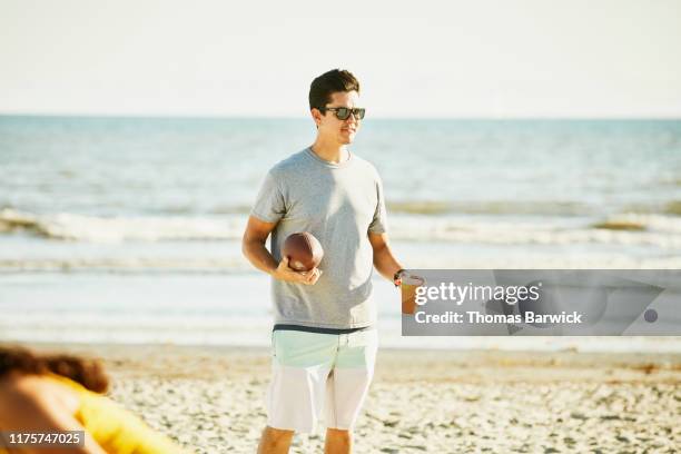 smiling man holding football and drink during party on beach - gray shorts stockfoto's en -beelden