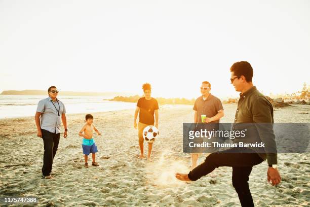 Man kicking soccer ball with friends on beach at sunset