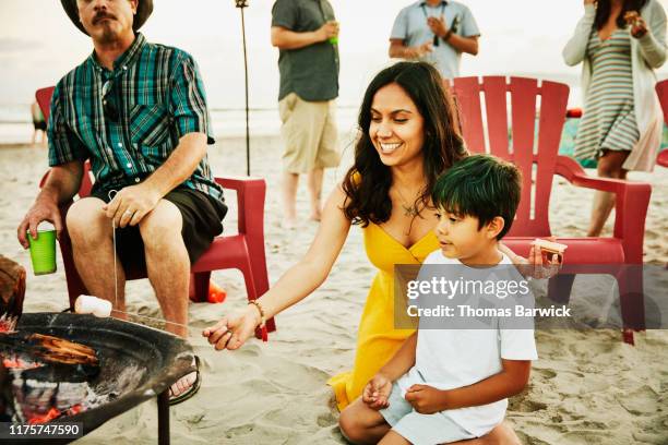 smiling aunt helping nephew roast marshmallows over fire during family beach party - aunyy stock pictures, royalty-free photos & images