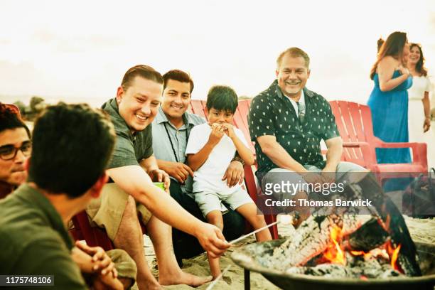 Laughing men hanging out by fire during family beach party
