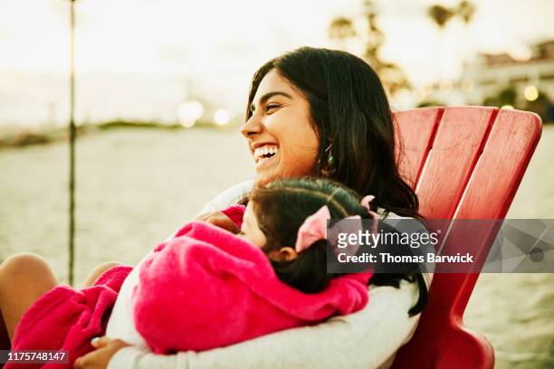 Laughing mother holding daughter wrapped in blanket in lap during family beach party