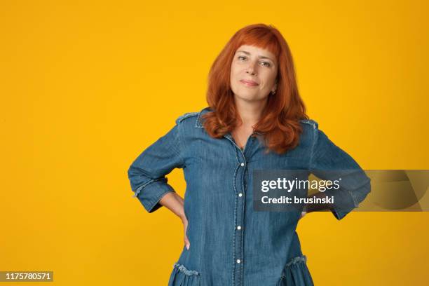 studioporträt einer 50-jährigen attraktiven rothaarigen frau in einem blauen kleid auf gelbem hintergrund - studio series stock-fotos und bilder