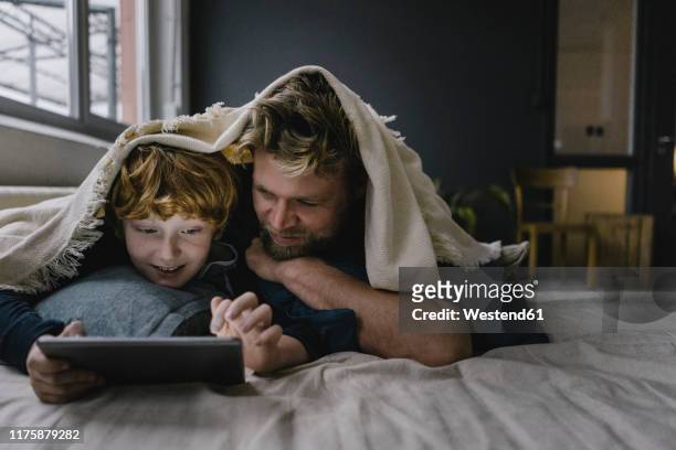 father and son lying together under blanket looking at digital tablet - future of media stock-fotos und bilder