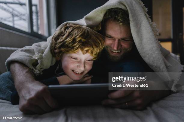 father and son lying together under blanket looking at digital tablet - dusk stock photos et images de collection