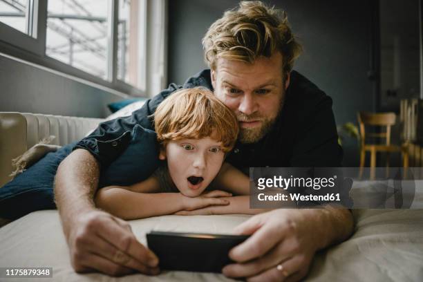 portrait of father and son lying on couch looking at cell phone - kind staunen stock-fotos und bilder