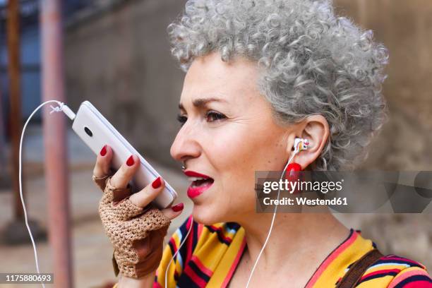 portrait of pierced mature woman with grey curly hair using earphones and cell phone - answering machine stock pictures, royalty-free photos & images