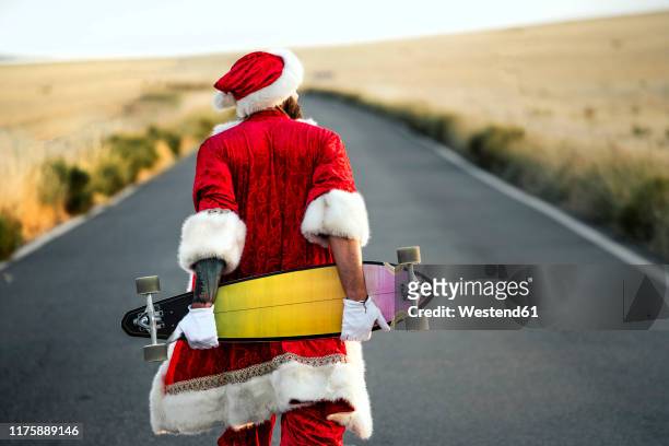 rear view of santa claus holding a longboard on country road - claus lange stock pictures, royalty-free photos & images