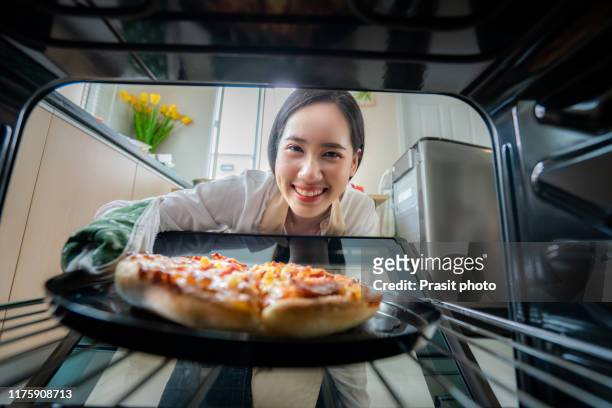 happy young asian woman looking at camera while cooking and put pizza in oven at kitchen at home . italian food, cooking, and people lifestyle concept. - backofen stock-fotos und bilder