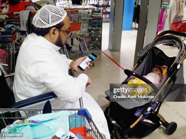 a middle eastern mature man using mobile phone and shopping in a mall on wheelchair with her grand daughter in a pram - saudi grandfather stock pictures, royalty-free photos & images