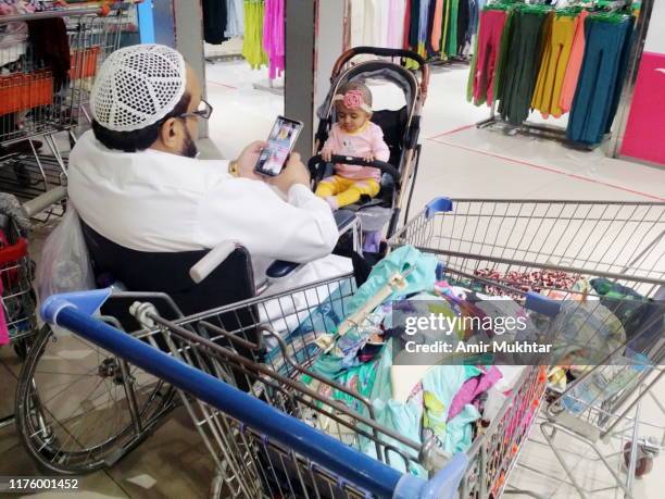 a middle eastern mature man using mobile phone and shopping in a mall on wheelchair with her grand daughter in a pram - saudi grandfather stock pictures, royalty-free photos & images