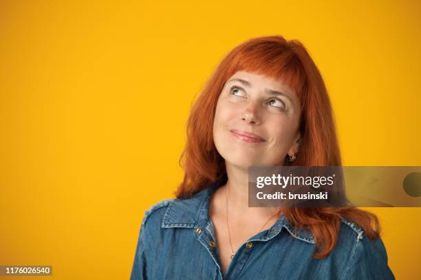 studio portrait of a 50 year old attractive red-haired woman in a blue dress on a yellow background - older woman colored hair stock pictures, royalty-free photos & images