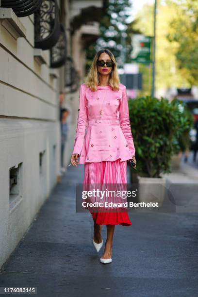 Candela Novembre wears sunglasses, a pink lustrous dress with attached rings and shoulder pads, a pink and red pleated skirt, white shoes, outside...