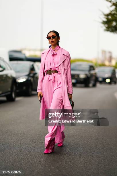 Gabriella Berdugo wears sunglasses, a pink trench coat, a pink flowing shirt, flared pants, pink shoes, a brown and pale green bag, a brown leather...