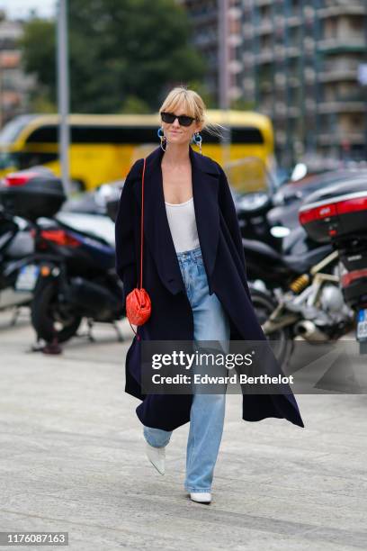 Jeanette Madsen wears sunglsses, blue earrings, a dark coat, a red bag, a white low neck top, blue flared denim jeans, white shoes, outside the...